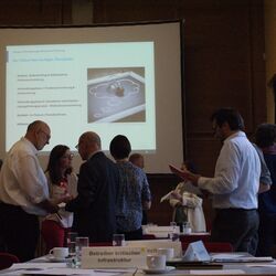 Ein Bild von dem Stakeholderdialog 2019: Katastrophenvorsorge bei Wasserengpässen kooperativ gestalten. Man sieht im Hintergund die PowerPoint Präsentation auf der das Planspiel erklärt ist, welches im Vodergrund abläuft. Personen stehen in kleineren Gruppen zusammen und diskutieren miteinander. 