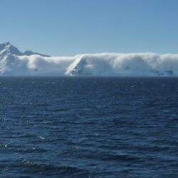 im Vordergrund des Bildes sieht man ruhiges antarktisches Meer. Im Hintergrund des Bildes schließen Berge an, die fast vollständig von Wolken eingehüllt sind. 