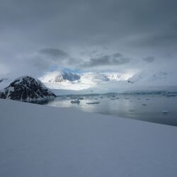 Im Vordergrund Schnee und Eis. Über dem Meer eine fast geschlossene Wolkendecke. Durch die Öffnung ist blauer Himmel zu sehen.