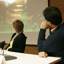Zwei Podiumsdiskutanten blicken auf eine Leinwand hinter sich, auf der ein Video gezeigt wird.