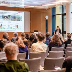 Teilnehmende folgen auf beigen Stühlen in Reihen sitzend einer Präsentation. Die Vortragende steht mit Handmikrofon nahe Stehpult vor einer Fensterfront.