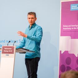 Vortragender in hellblauem Hemd steht gestikulierend am Stehpult vor dem blauen Hintergrund, auf dem der Veranstaltungsort geschrieben steht. Daneben steht das lilafarbene Veranstaltungs-Rollup.