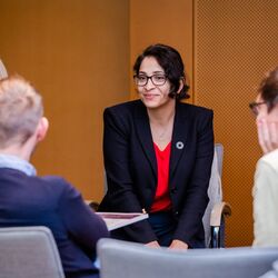 Teilnehmende sitzen in aufgelockerter Stimmung beieinander im holzfarben vertäfelten Veranstaltungsraum. Die Teilnehmerin im Mittelpunkt trägt ein dunkelblaues Sacko mit rotes Oberteil.