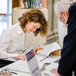Ein Teilnehmender registriert sich am Registriertisch und trägt sich in die Teilnehmendenliste ein. Eine Dame in einer weißen Bluse trägt Angaben in eine Liste ein.