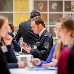 Teilnehmende stehen in der Kaffeepause an Stehtischen zusammen und tauschen sich rege aus. Ein uniformierter Teilnehmender isst Kuchen.