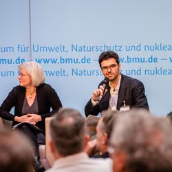 Podiumsdiskussion vor dem hellblauem Hintergrund, auf dem der Veranstaltungsort geschrieben steht. Ein Teilnehmender hält ein Handmikrofon.