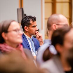 Der Blick geht durch die Teilnehmenden auf einen Teilnehmer, der sehr nachdenklich schaut. Er trägt einen blauen Anzug mit hellblauem Schlips und weisses Hemd. Der Raum ist hell und holzfarben abgesetzt.