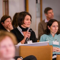 Teilnehmende sitzen im einem Veranstaltungsraum auf orangenen Stühlen in Reihen und blicken nachdenklich. Eine Teilnehmerin spricht in ein Handmikro.