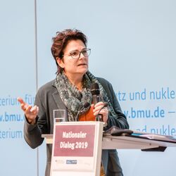 Zwei Vortragende stehen am Stehpult vor blauem Hintergrund, auf dem der Veranstaltungsort geschrieben steht. Beide halten Handmikrofone in den Händen und sprechen gestikulierend  miteinander.