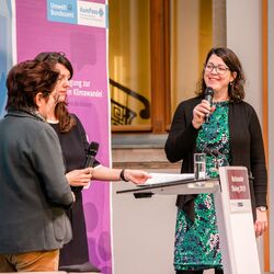 Drei Vortragende stehen am Stehpult und halten Mikrofone in den Händen. Sie lachen sich gegenseitig an.Im Hintergrund steht das lilafarbene Veranstaltungs-Rollup