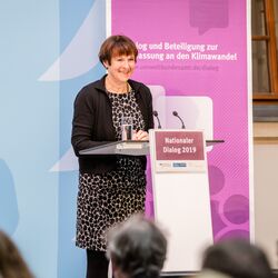 Teilnehmende hält eine Rede am Stehpult. Auf dem Stehpult steht ein Glas Wasser. Im Hintergrund steht ein Aufsteller sowie vorne auf dem Stehpult mit dem Veranstaltungstitel vorwiegend in lila gehalten.