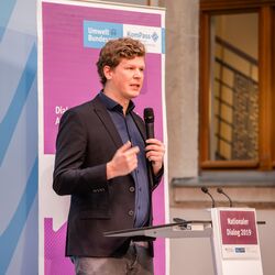 Vortragender steht mit Handmikrofon am Stehpult vor dem lilafarbenen Veranstaltungs-Rollup. Im Hintergrund geht der Blick durch ein hölzernes Fensterkreuz in den Flur des Gebäudes.