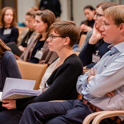 Teilnehmende sitzen im Veranstaltungsraum auf orangenen Stühle in Reihen und schauen zuhörend in eine Richtung.