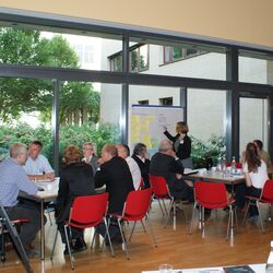 Teilnehmer sitzen an Gruppenarbeitstischen im Raum vor einer großen Fensterfront. Eine Teilnehmerin schreibt im Hintergrund Text auf Papier an Moderationswand.