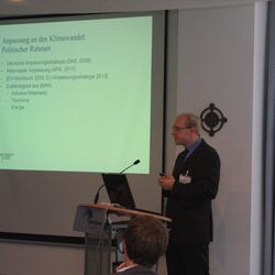 Vortragender steht am Rednerpult. Im Hintergrund ist die Präsentation auf Projektionswand zu sehen und der Veranstaltungsaufsteller.