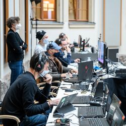 Die Technikerinnen und Techniker sitzen in einer langen Reihe und schauen konzentriert in die Laptops. Sie tragen Kopfhörer und Mund-/Nasenbedeckungen.