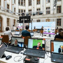 In dem hellen historischen Innenhof geht der Blick über den langen Tisch mit dem Laptops hinüber über sitzendes Publikum zum Podium. Auf dem Podium sitzen entfernt zwei Menschen vor blauem Hintergrund. Im Hof stehen verteilt auf Ständern die flachen Bildschirme, die die dazugeschalteten Teilnehmenden zeigen. Kamera- und Lichttechnik hängt  in der Höhe seitlich entlang der Fassade an Metallstreben.