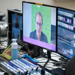 Der Blick geht auf den Technikerinnentisch Mischpult und Computerbildschirmen. Darauf sind die Teilnehmenden und verschiedenen Konferenzräume zu sehen.