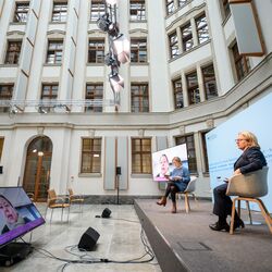 In einem historischen Innenhof mit natürlichen Licht sitzen zwei Frauen auf einem Podium auf hellen Schalenstühlen vor hellblauem Hintergrund und schauen vor sich runter auf einen flachen Bildschirm. Vor dem flachen Bildschirm stehen zwei schwarze Lautsprecher auf dem Boden. Im Hintergrund sitzt ein Mann auf einem Publikumsstuhl. Die Fassaden sind in einem freundlichen Hellgrau abgesetzt, die Fenster sind hölzern.