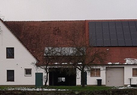 Ein großes Bauernhaus mit Solarkollektoren und Photovoltaik auf dem Dach vor teils verschneiter Wiese mit kahlen Obstbäumen