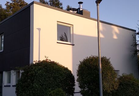 Ein zweigeschossiges, weißes Einfamilienhaus mit Flachdach, schräg von der Straße gesehen; das Obergeschoss über dem Eingang ist mit dunklem Holz verkleidet