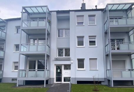 Ein dreistöckges, hellgraues Mehrfamilienhaus mit links und rechts vom Eingang vorangestellten Balkonen, frontal von vorne gesehen