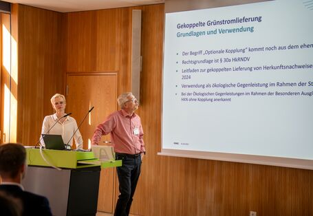 Eine Person referiert mit Blick auf eine projizierte Präsentation. Eine weitere Person steht daneben. Im Vordergrund ist unscharf ein Hinterkopf abgebildet.