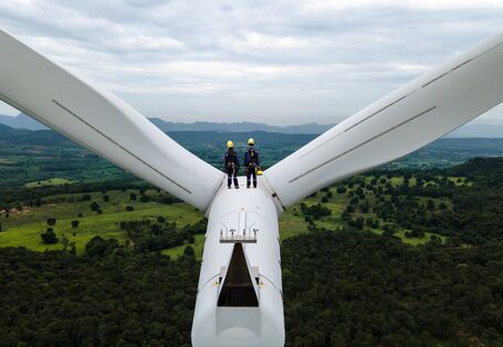 Zwei Menschen i9n Arbeitskleidung stehen auf einer Winkraftanlage und schauen in einer bewaldete Landschaft.
