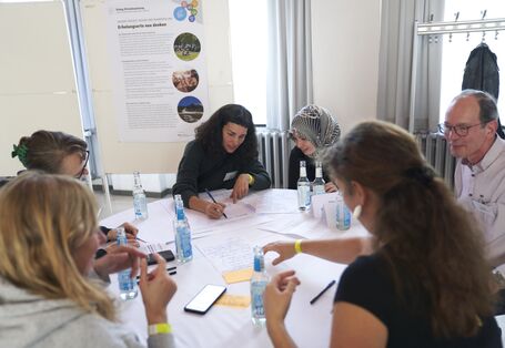 Das Foto zeigt einen Tisch mit mehreren Menschen, von denen eine Teilnehmerin Notizen macht.