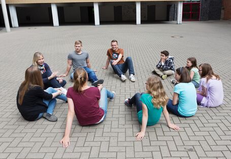 Auf dem Bild sind Jugendliche zusehen, die in einem Kreis auf dem Schulhof sitzen.