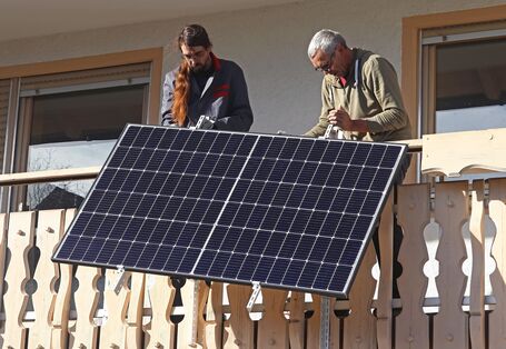 Zwei Personen installieren ein Solarpanel an Holzgeländer eines Gebäudes, im Hintergrund sind Fenster zu sehen.
