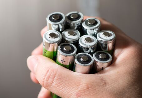 Eine Hand hält ein Bündel gebrauchter AA-Batterien fest umschlossen. Die Pole der Batterien zeigen nach oben, das Gehäuse einiger Batterien ist grün-silberfarben.
