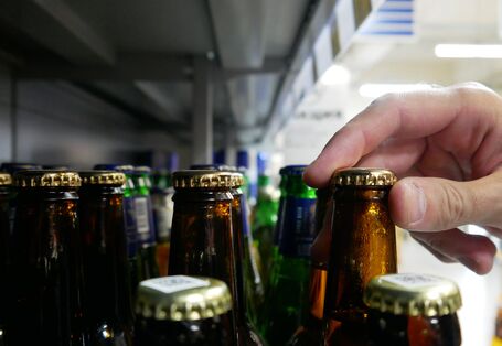 Eine Hand greift in einem Supermarktregal nach dem Kronkorken einer braunen Glasflasche, die zwischen anderen braunen und grünen Flaschen steht.