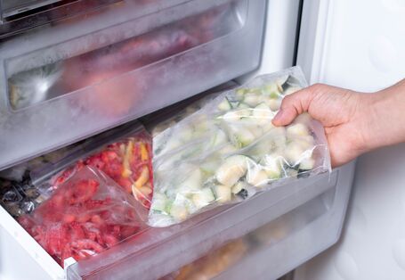 Eine Hand holt eine Plastiktüte mit gefrorenen Zucchini-Stücken aus dem Gefrierfach eines Kühlschranks. Im Fach darunter befinden sich Tüten mit anderen gefrorenen Lebensmitteln, wie zum Beispiel Paprika.