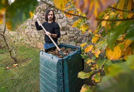 Ein Mann mit dunklen Haaren und Bart in einem schwarzen Pullover und blauen Jeans steht im Garten vor einer grünen Komposttonne und mischt den Inhalt mit einem Rechen um. Im Hintergrund ist eine Natursteinmauer zu sehen und im Vordergrund hängt ein Ast mit gelben und grünen Blättern.