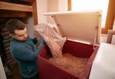 Ein Mann mit einem blauen Pullover leert einen durchsichtigen Sack mit Holzpellets in einen roten Behälter, der zu einer Pelletheizung gehört. Im Hintergrund ist ein Fenster zu sehen, durch das Sonnenlicht in den Raum fällt.