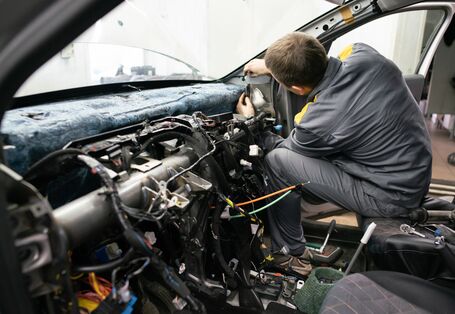 Ein Mechaniker in grauer Arbeitskleidung sitzt auf dem Beifahrersitz eines Autos und arbeitet am zerlegten Armaturenbrett. Das Innere des Fahrzeugs ist offen gelegt, wobei zahlreiche Kabel, Steckverbindungen und die Metallstruktur der Instrumententafel sichtbar sind.