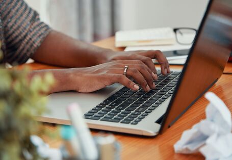 Eine Person mit einem silbernen Ring an der Hand tippt auf einer Laptoptastatur. Im Hintergrund sind verschwommen eine Brille, ein Stift und zerknülltes Papier zu sehen. 