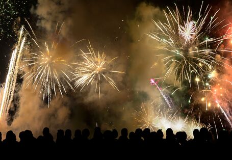 Silhouetten einer großen Menschenmenge, die vor einem dunklen Nachthimmel ein großes, farbenfrohes Feuerwerk mit goldenen und rötlichen Lichteffekten sowie Rauchwolken betrachtet.