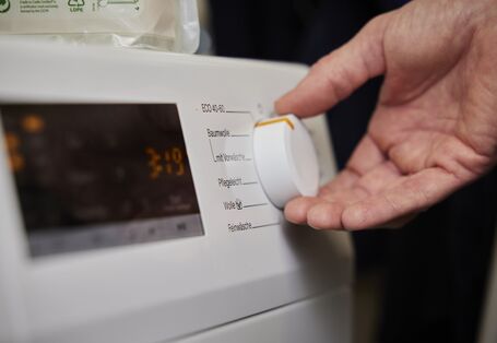 Eine Nahaufnahme zeigt eine Hand, die den Drehknopf einer Waschmaschine auf ein Programm einstellt. Auf dem Bedienfeld der Maschine sind verschiedene Waschprogramme wie „ECO 40-60“, „Baumwolle“ und „Pflegeleicht“ zu sehen.