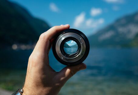 A hand holding a lens can be seen in the foreground. A blurred mountain lake landscape can be seen in the background, the lens focuses into the horizon.
