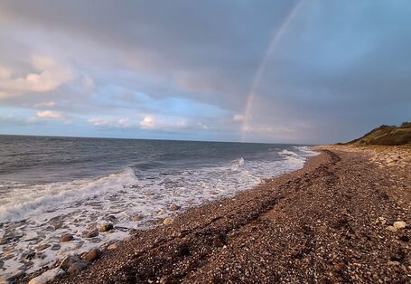 Wellen der Ostsee schlagen an einen Steinstrand, der in grüne Hügel übergeht. Der Himmel ist über den Hügeln graublau aber über dem Meer hellblau mit weißen Wolken. Ein Regenbogen beginnt über dem Meer.  