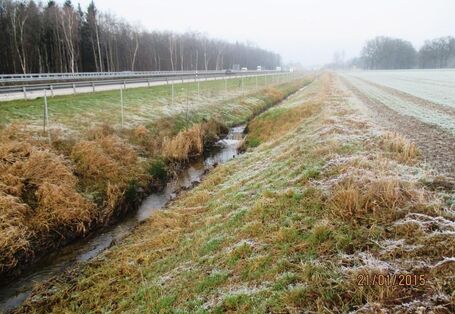 Foto: Der begradigte Schierenbach im V-Profil eingeengt zwischen Autobahn und Ackerfläche. Die Ufer sind bis auf kurze Gräser frei von Vegetation.