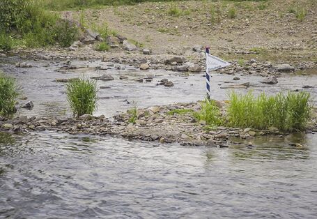 Foto: Eine bewachsene Kiesbank mit Wimpel der Schützenkompanie „Schütten – Dinschede“ an einer kleinen Fahnenstange.