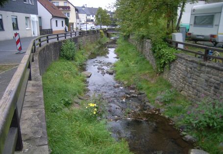 Foto: Ein Bach mit hohen Ufermauern und angrenzenden Häusern und Straßen in Ortslage. Die Gewässersohle ist offen, mit Kies bedeckt und stellenweise bewachsen. Der Bach verläuft leicht schwingend im Kastenprofil.
