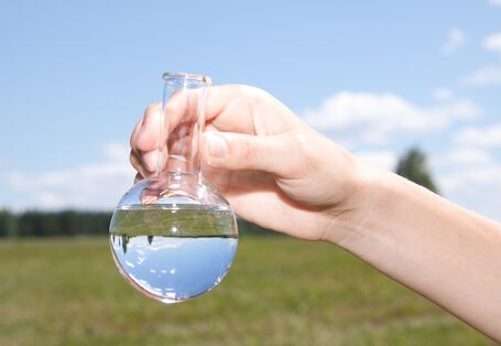 Ein Reagenzkolben vor grüner Wiese, darin Wasser.
