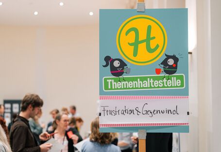 Auf einer Themenhaltestelle ist das Thema "Frustration und Gegenwind" angebacht und Teilnehmende stehen in der Nähe uns tauschen sich aus