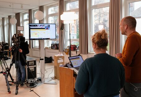 Zwei Moderatoren gucken in die Kamera und auf einen Bildschirm, während Sie das Netzwerktreffen moderieren.