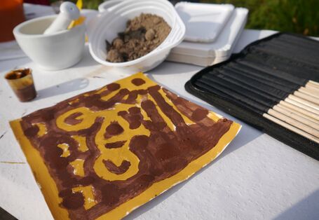 Dargestellt ist ein Fahrrad im Sonnenschein, gemalt mit Bodenfarben von Teilnehmenden der Fortbildung, im Hintergrund der verwendete Boden in einer Schale