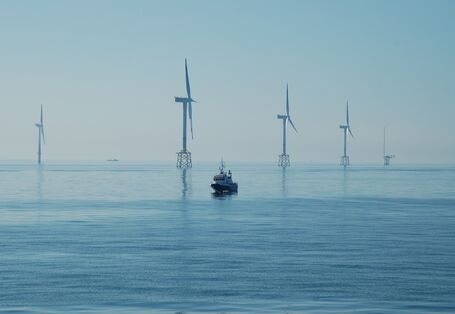 Das Bild zeigt im Meer stehende Windräder eines Offshore-Windparks und ein dazwischen schwimmendes Schiff.